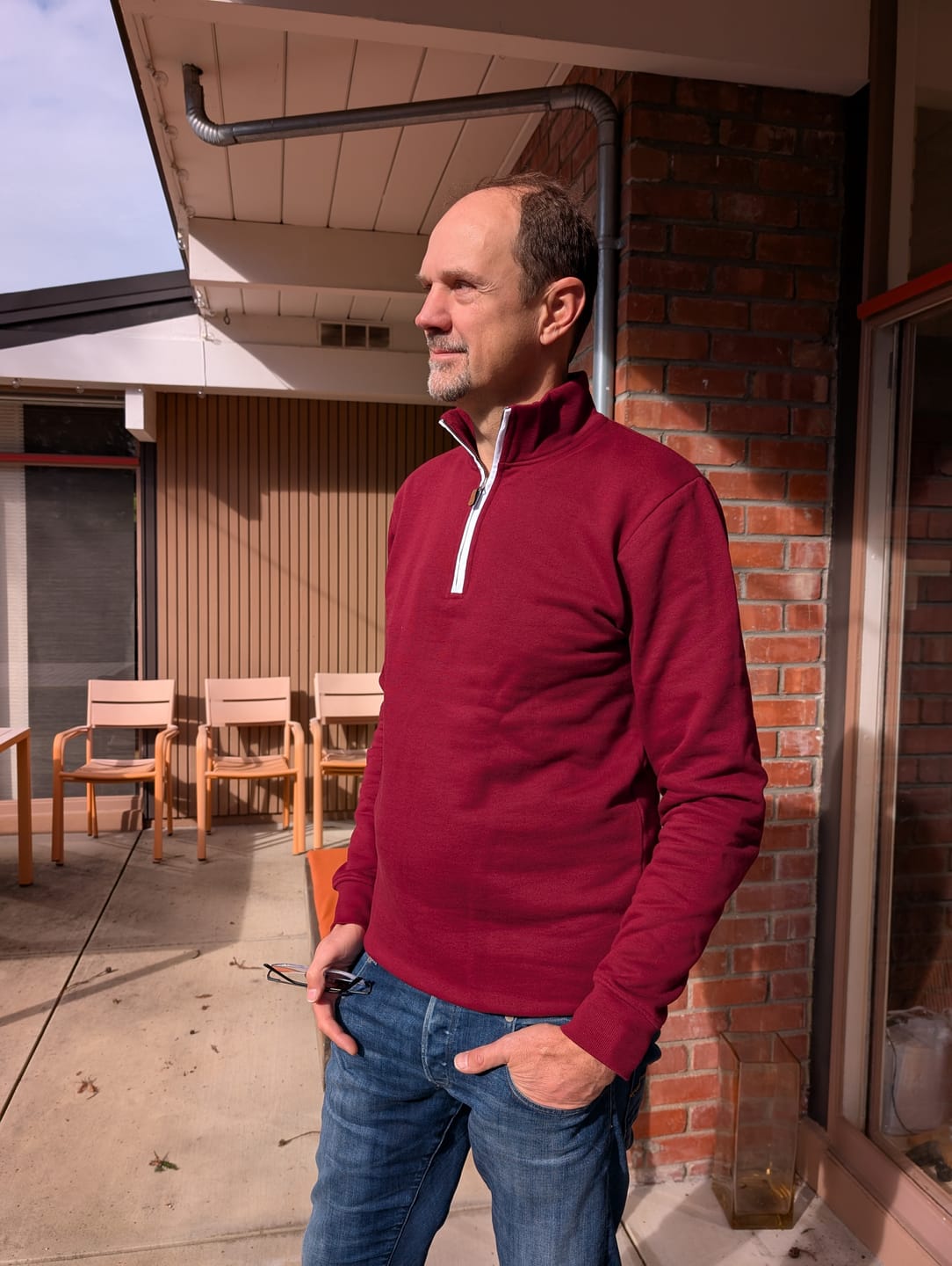 Tall man wearing a maroon tall quarter-zip sweatshirt and standing outside a building with chairs in the background.