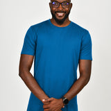 A tall and slim man in the studio standing in front of a light background. The smiling model is wearing an extra long slim azure t-shirt in a size large. The tall large t-shirt features a 3" longer body, 100% organic cotton, and is soft & preshrunk. The large t-shirt is ideal for tall slim men 6'2"+.