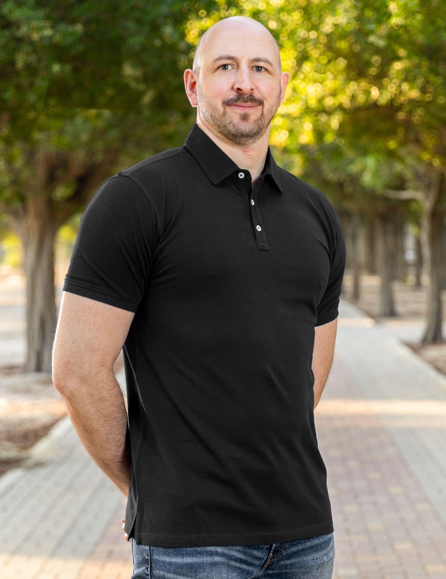 A tall and athletic man in a park standing on a path between trees. The smiling model is wearing an extra long slim black polo shirt in a size XL. The tall black polo shirt features a 3" longer body, 100% organic cotton, and is soft & preshrunk. The black polo shirt is ideal for tall slim men 6'2"+.