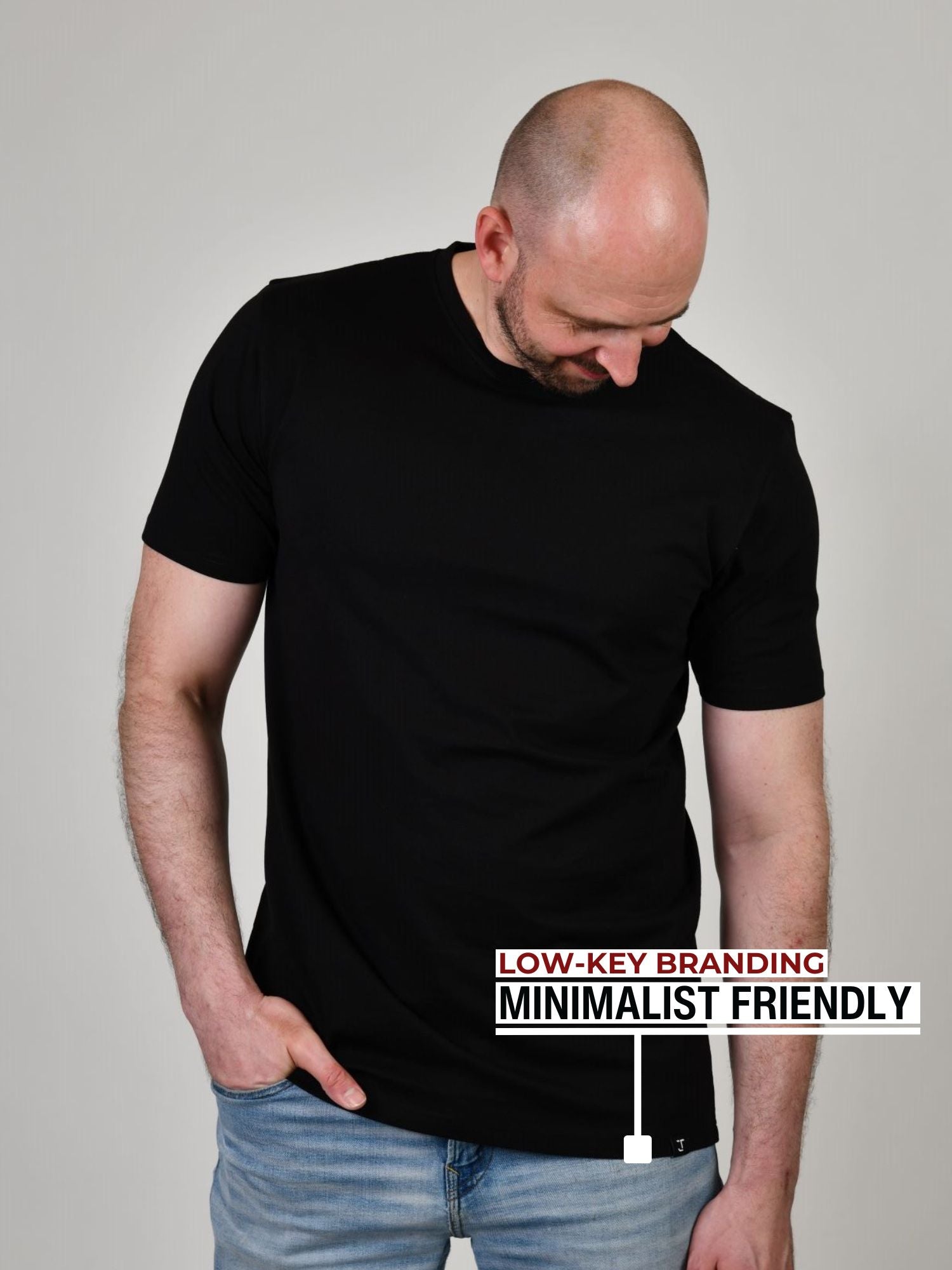 A tall athletic guy standing in a studio and wearing a black XL tall t-shirt, looking down at the Just Tall monogram logo.