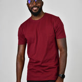 A tall and slim man in the studio standing in front of a light background. The smiling model is wearing an extra long slim cabernet t-shirt in a size large. The tall cabernet t-shirt features a 3" longer body, 100% organic cotton, and is soft & preshrunk. The cabernet t-shirt is ideal for tall slim men 6'2"+.
