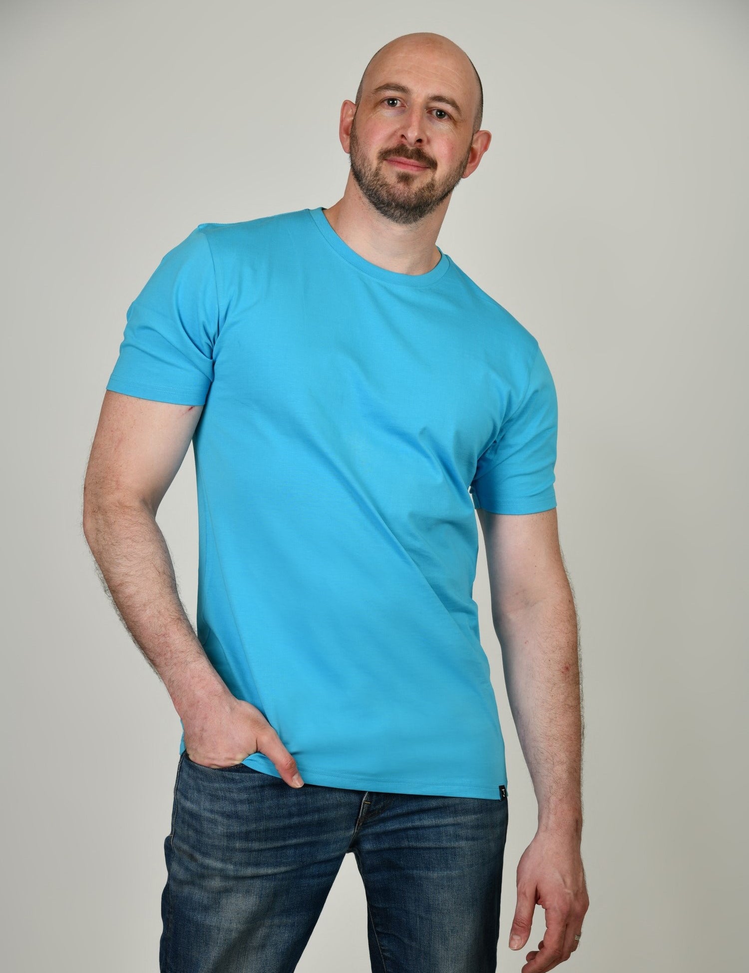 A tall and slim man in the studio standing in front of a light background. The smiling model is wearing an extra long slim cyan t-shirt in a size XL. The tall cyan t-shirt features a 3" longer body, 100% organic cotton, and is soft & preshrunk. The cyan t-shirt is ideal for tall slim men 6'2"+.