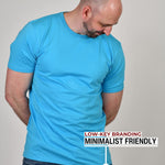 An upper body shot of a tall athletic guy in a studio and wearing a cyan extra large tall slim t-shirt and looking down at the monogram logo.