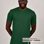 A tall slim guy standing in a studio and wearing a dark green large tall t-shirt, smiling and hands behind back.