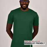 A tall slim guy standing in a studio and wearing a dark green large tall t-shirt, smiling and hands behind back.