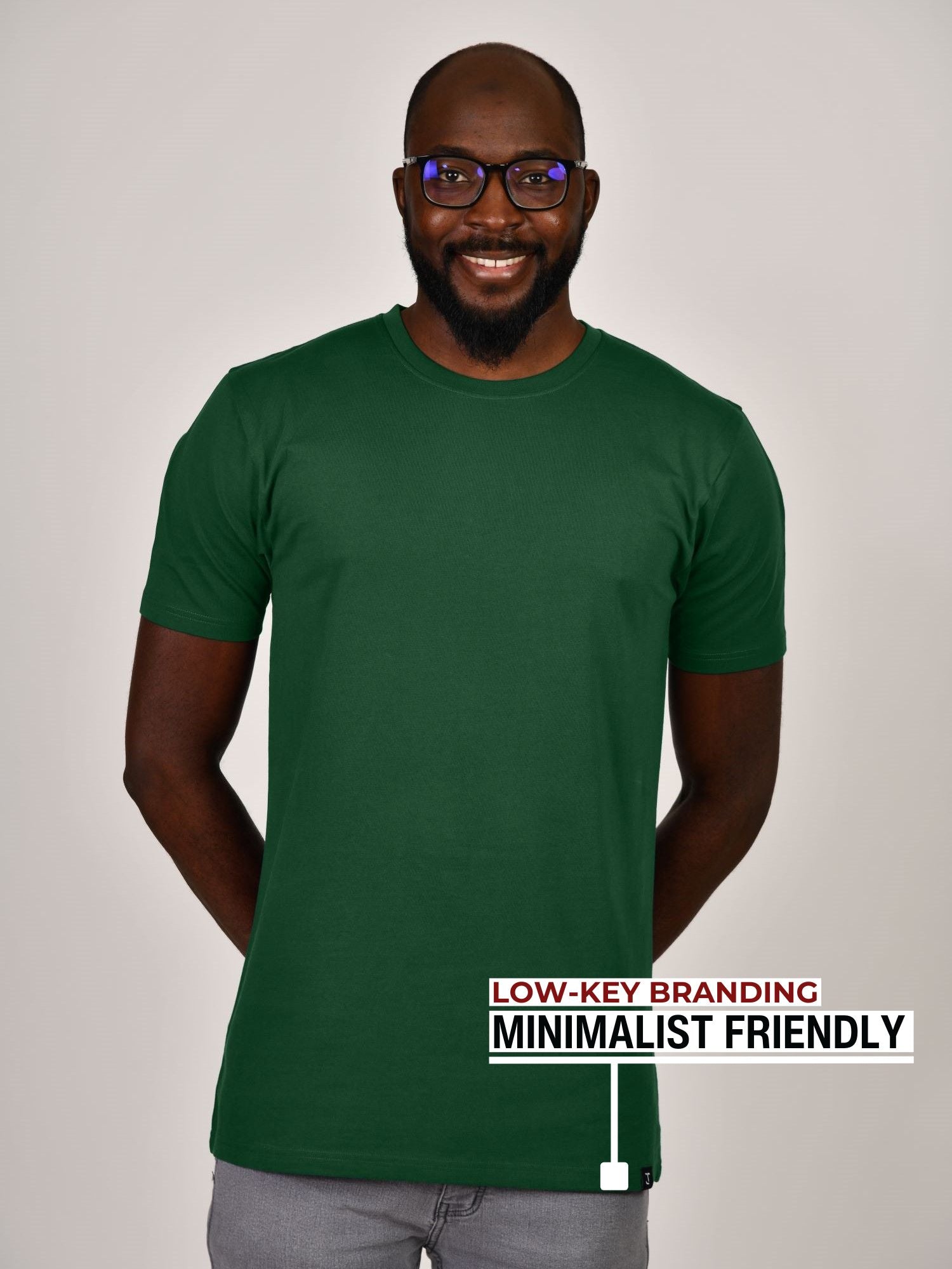 A tall slim guy standing in a studio and wearing a dark green large tall t-shirt, smiling and hands behind back.