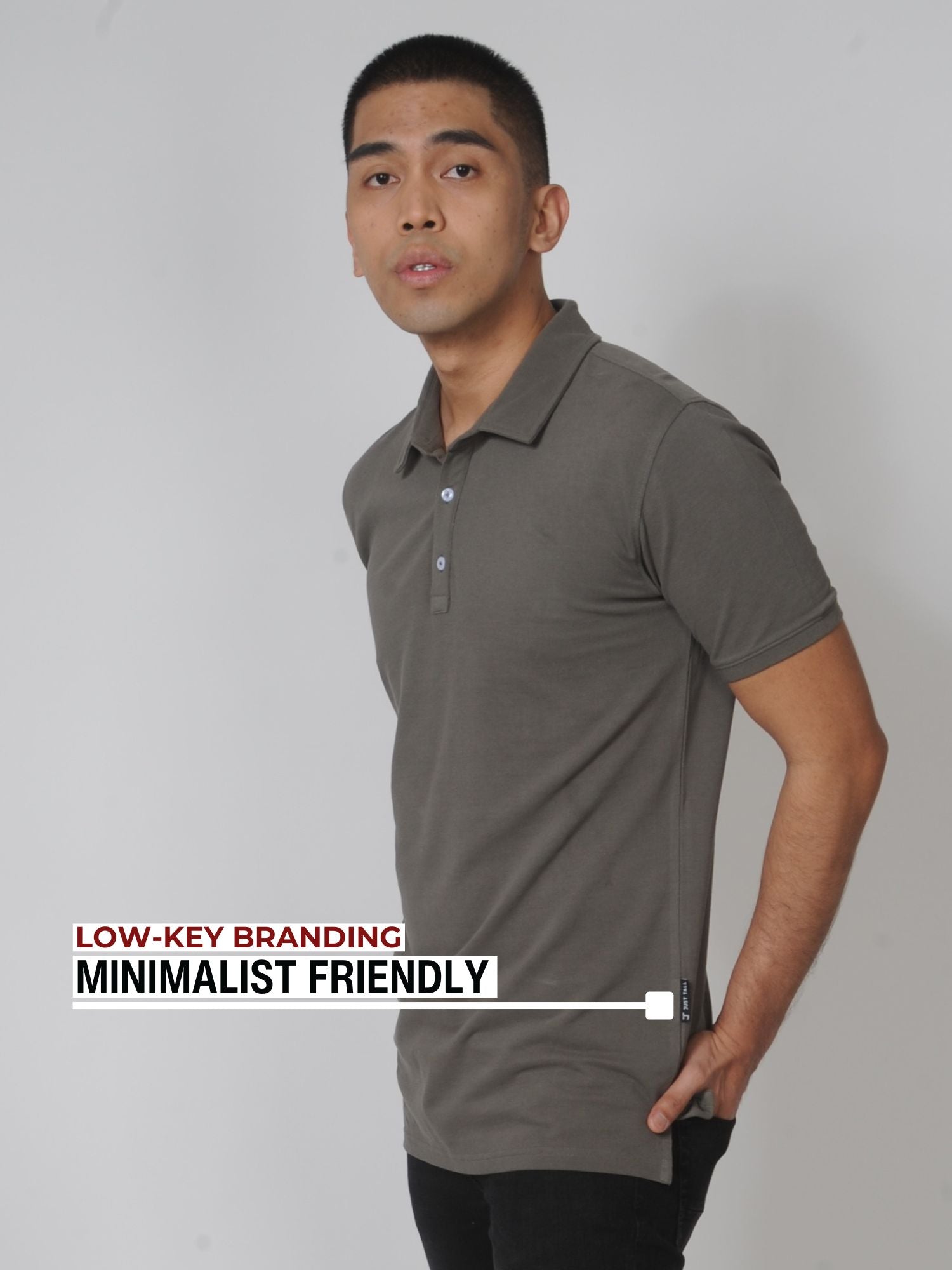 An upper body shot of a tall slim guy in a studio and wearing a medium tall slim dark grey polo shirt and showing the minimal Just Tall flag logo near the bottom of the side seam.