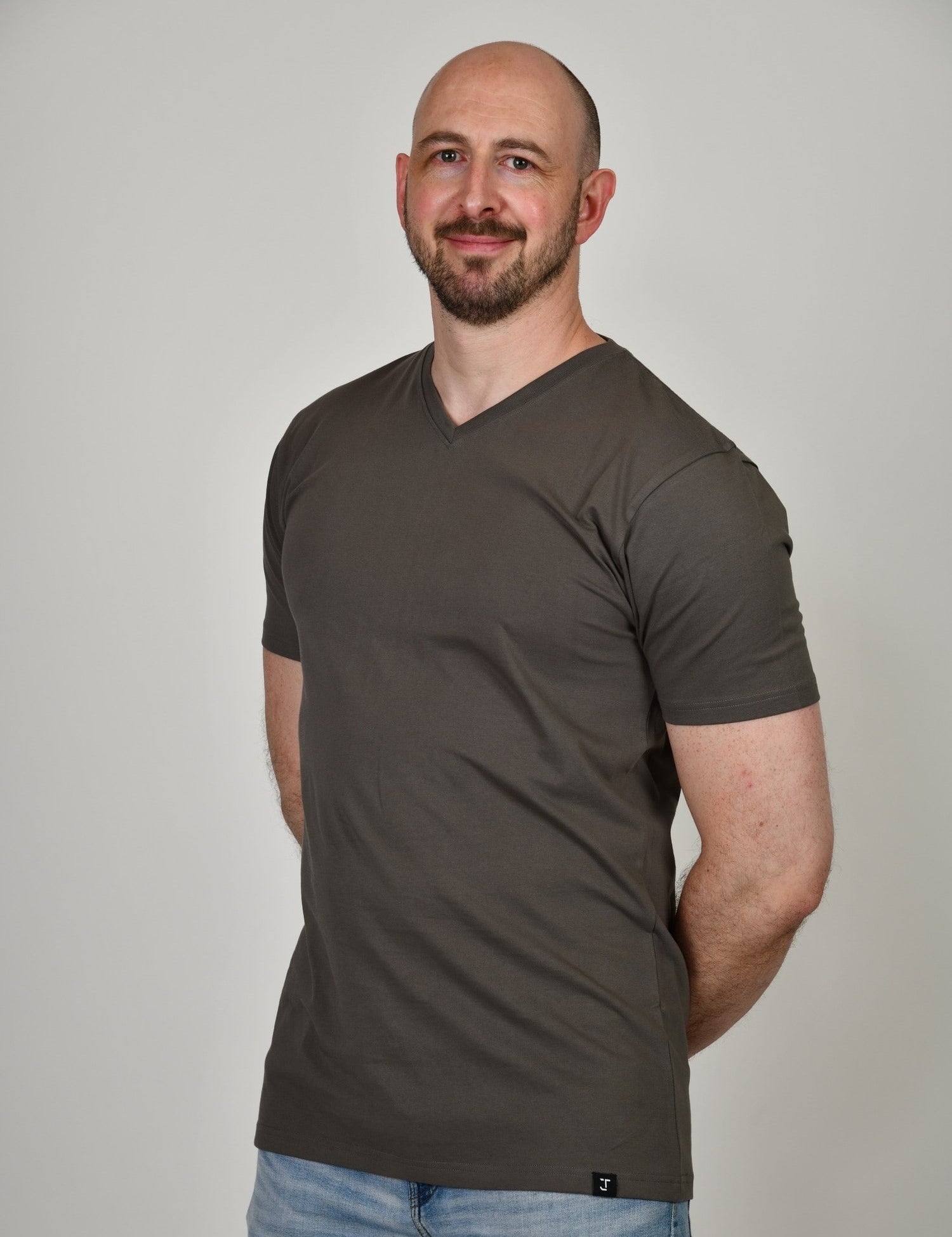 A tall and slim man in the studio standing in front of a light background with hands behind back. The smiling model is wearing an extra long slim dark grey v-neck t-shirt in a size XL. The tall dark grey v-neck t-shirt features a 3" longer body, 100% organic cotton, and is soft & preshrunk. The dark grey v-neck t-shirt is ideal for tall slim men 6'2"+.