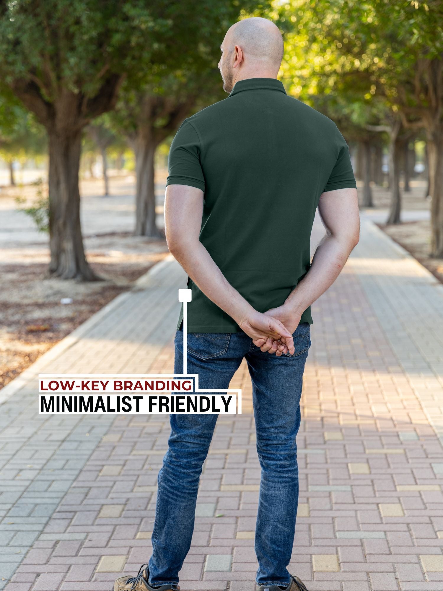 A shot from behind of a tall athletic guy in a park and wearing an XL tall slim dark pine polo shirt and showing the minimal Just Tall flag logo near the bottom of the side seam.