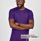 A tall slim guy standing in a studio and wearing a royal purple large tall t-shirt, smiling and arms folded.