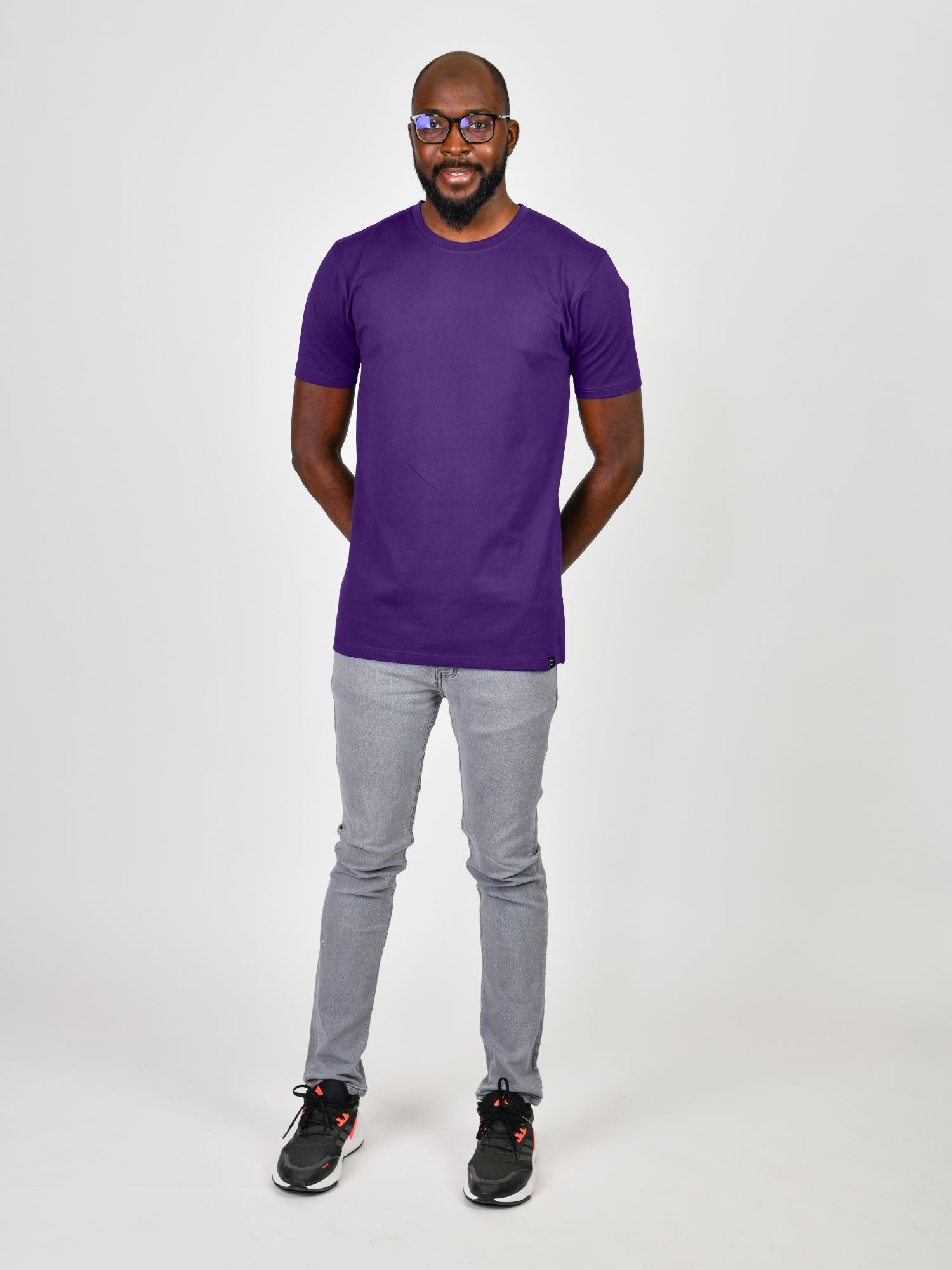 A head to toe shot of a tall slim guy in a studio and wearing a royal purple large tall slim t-shirt, hands behind back.