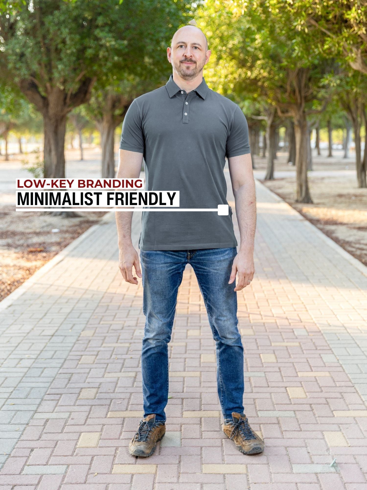 A head to toe shot of a tall athletic guy in a park and wearing an XL tall slim dark slate polo shirt and showing the minimal Just Tall flag logo near the bottom of the side seam.
