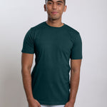 A tall and slim man in the studio standing in front of a light background. The smiling model is wearing an extra long slim dark teal t-shirt in a size medium. The tall dark teal t-shirt features a 3" longer body, 100% organic cotton, and is soft & preshrunk. The dark teal t-shirt is ideal for tall slim men 6'2"+.