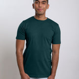 A tall and slim man in the studio standing in front of a light background. The smiling model is wearing an extra long slim dark teal t-shirt in a size medium. The tall dark teal t-shirt features a 3" longer body, 100% organic cotton, and is soft & preshrunk. The dark teal t-shirt is ideal for tall slim men 6'2"+.
