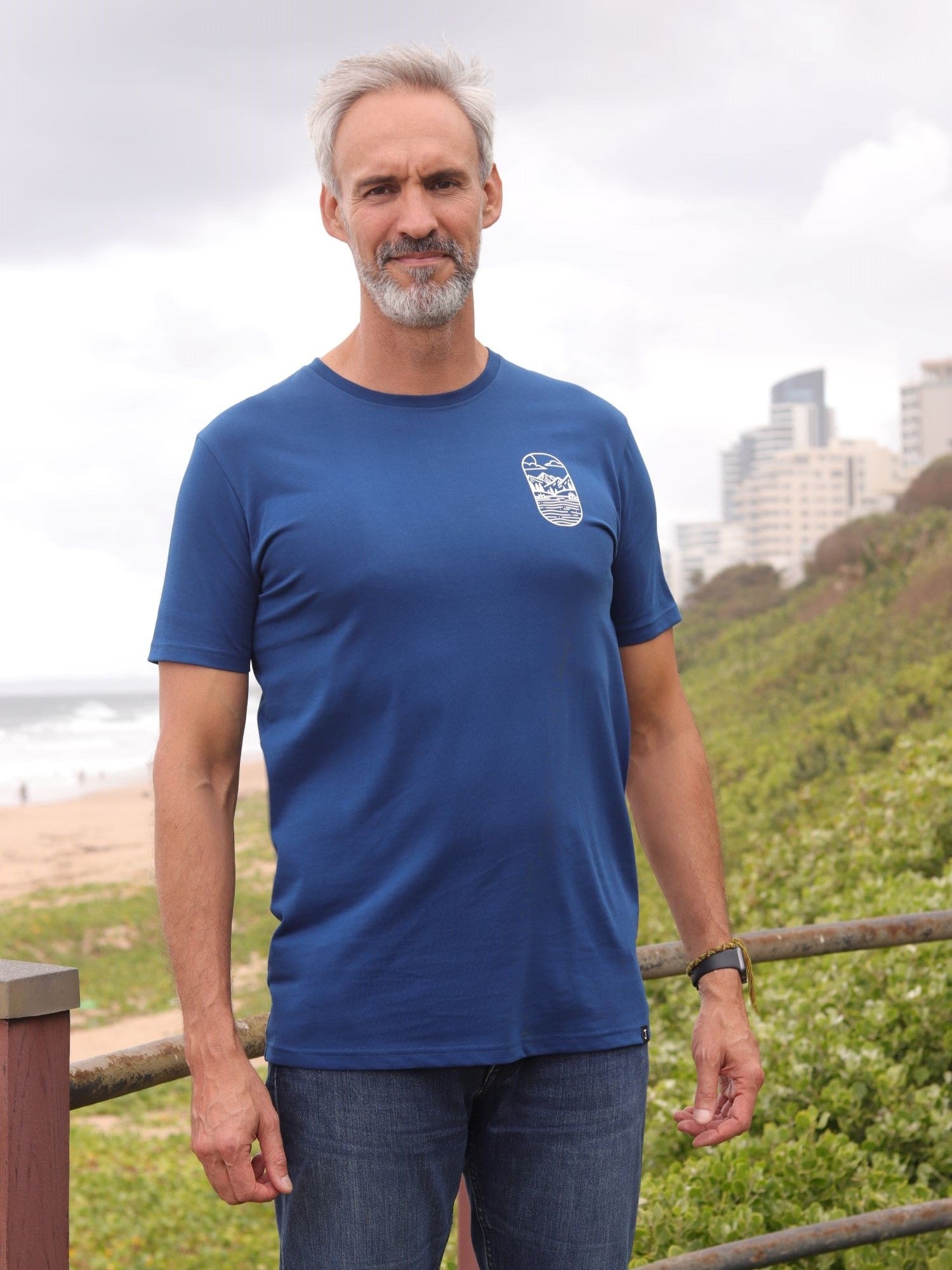 A tall and slim man standing by the sea. The smiling model is wearing a navy blue extra long graphic t-shirt with a minimal lake design. It features a 3" longer body, 100% organic cotton, and is soft & preshrunk. This extra long tall graphic tee is ideal for tall slim men 6'2"+