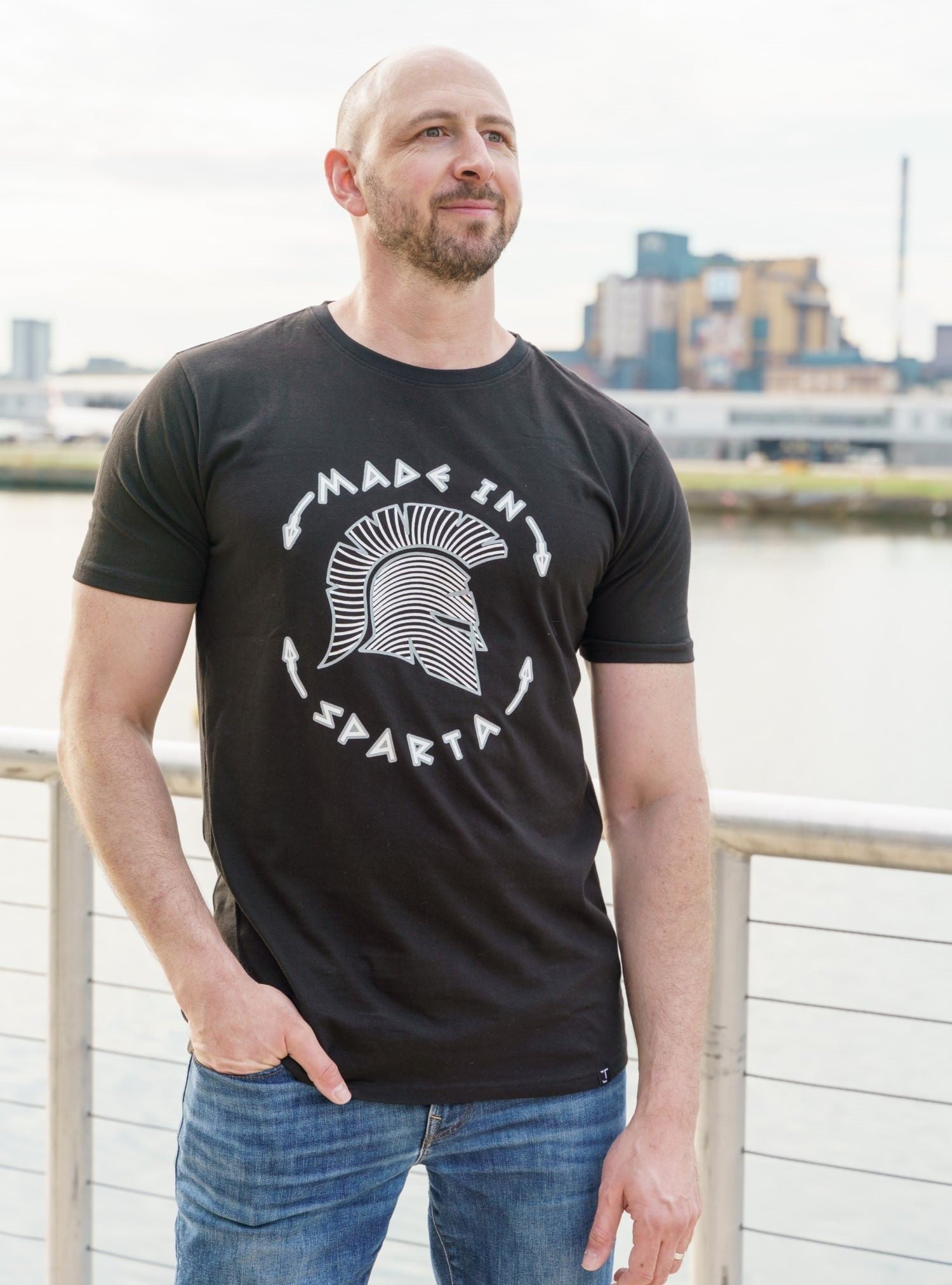 A tall and slim man standing by a canal. The smiling model is wearing a black extra long graphic t-shirt with the text made in sparta and a spartan helmet. It features a 3" longer body, 100% organic cotton, and is soft & preshrunk. This extra long tall graphic tee is ideal for tall slim men 6'2"+.