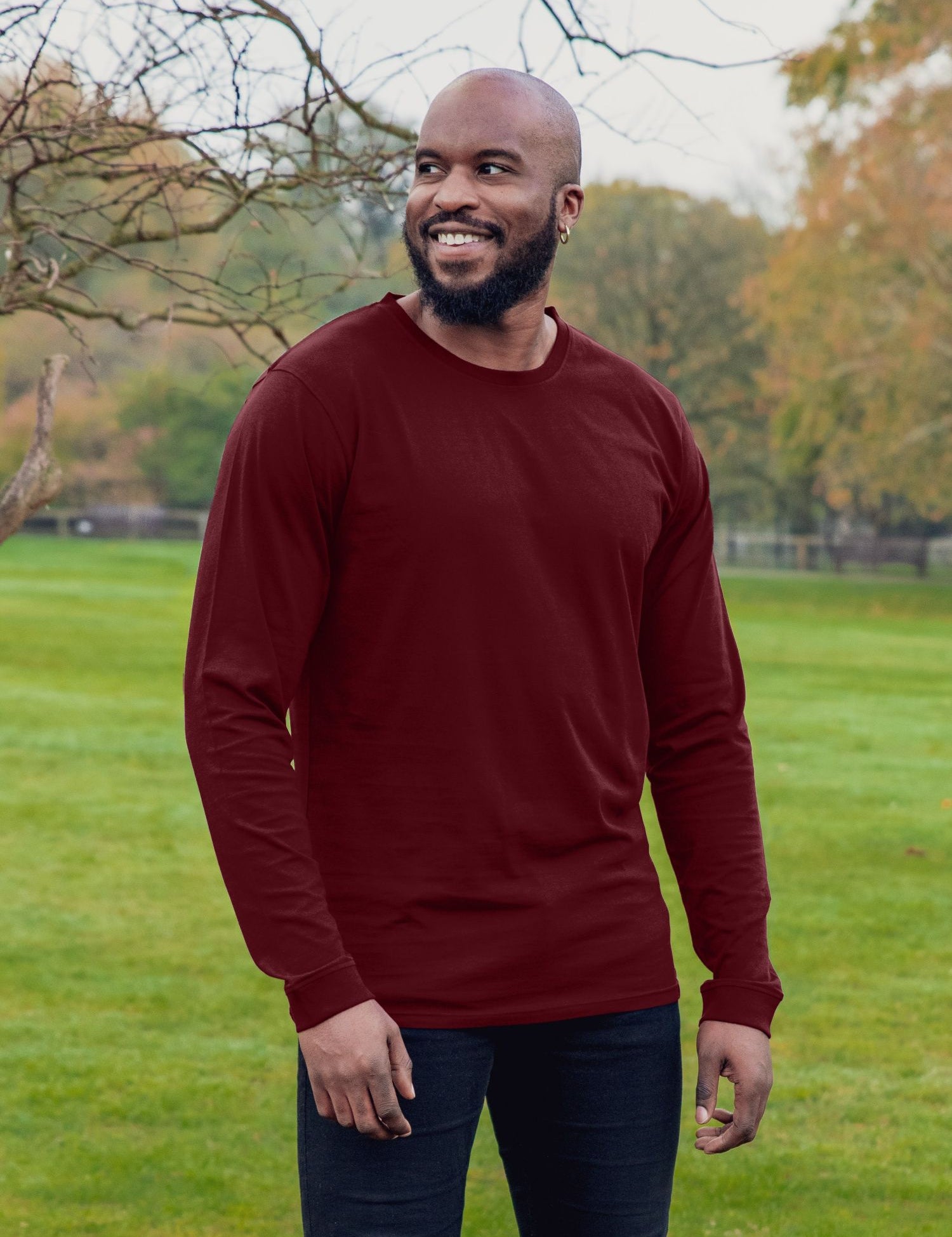 An upper body shot of a tall athletic guy wearing an XL tall maroon long sleeve t-shirt, standing in a park and smiling.