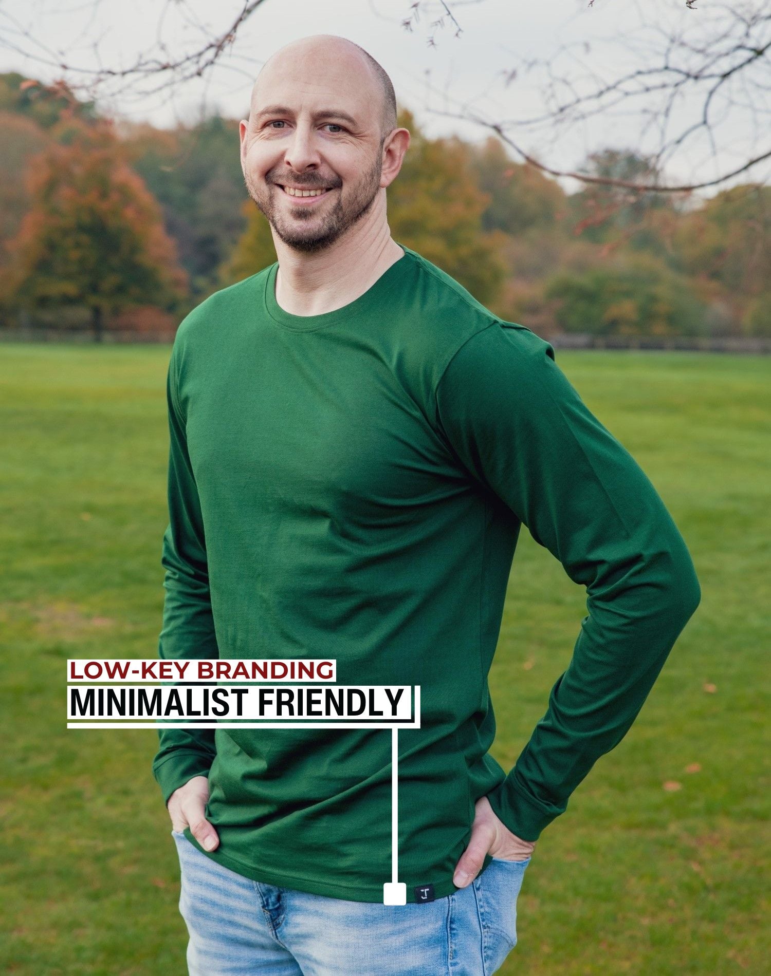 An upper body shot of a tall athletic guy standing in a park wearing a dark green extra long tall long sleeve t-shirt and showing the Just Tall minimalist monogram logo.