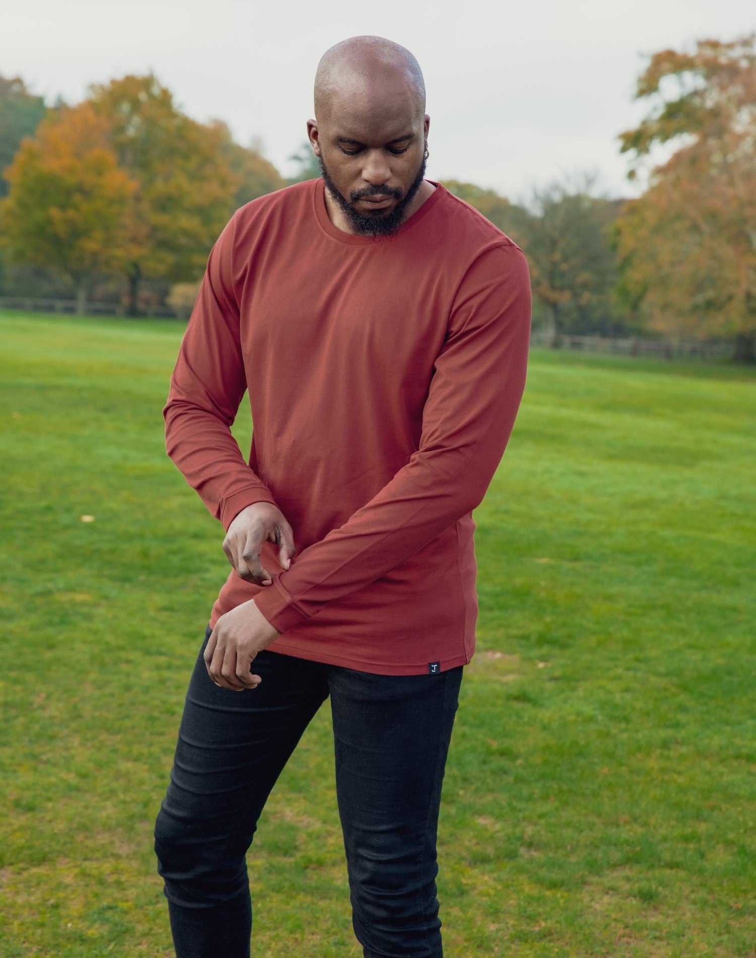 A shot of a tall athletic guy in a park wearing a dark orange long sleeve tall t-shirt, looking down at the t-shirt logo.