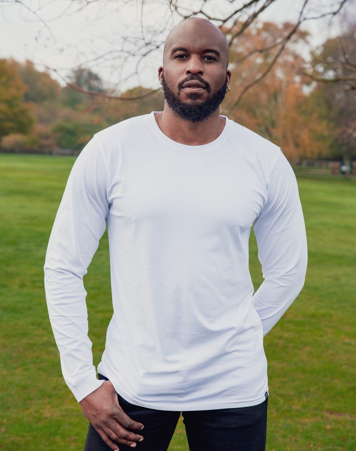 A tall and slim man standing in a park with one hand in his pocket. The smiling model is wearing a white long-sleeve tall t-shirt. The tall white long-sleeve t-shirt features a 2.5" longer sleeves, 100% organic cotton, and is soft & preshrunk. The white long-sleeve t-shirt is ideal for tall slim men 6'2"+.