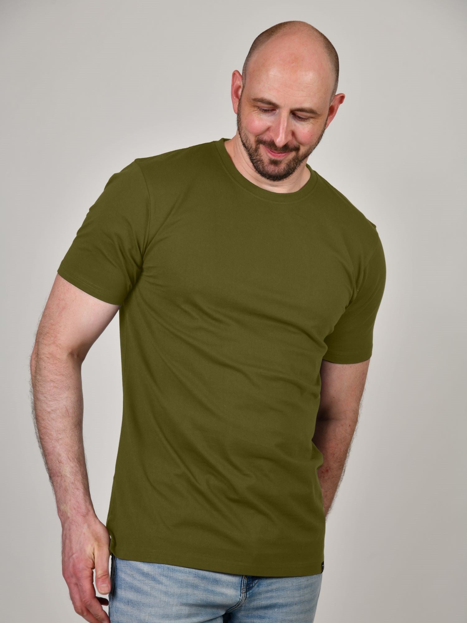 A tall and slim man in the studio standing in front of a light background. The smiling model is wearing an extra long slim olive t-shirt in a size XL. The tall olive t-shirt features a 3" longer body, 100% organic cotton, and is soft & preshrunk. The olive t-shirt is ideal for tall slim men 6'2"+.