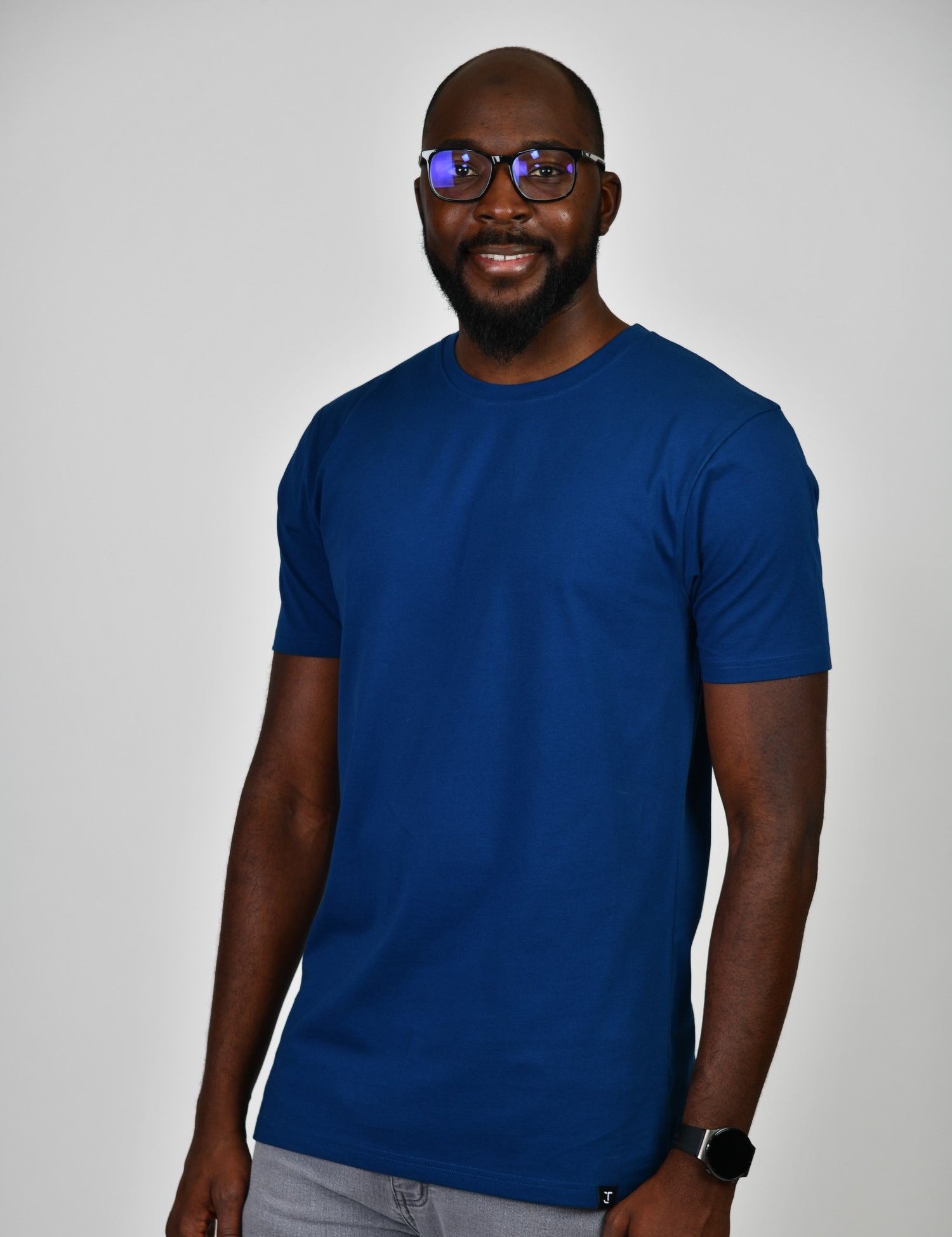 A tall and slim man in the studio standing in front of a light background. The smiling model is wearing an extra long slim navy t-shirt in a size large. The tall navy t-shirt features a 3" longer body, 100% organic cotton, and is soft & preshrunk. The navy t-shirt is ideal for tall slim men 6'2"+.