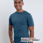 An upper body shot of a tall slim guy in a studio and wearing a petrol medium tall t-shirt, smiling and hand in pocket.