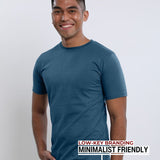 An upper body shot of a tall slim guy in a studio and wearing a petrol medium tall t-shirt, smiling and hand in pocket.