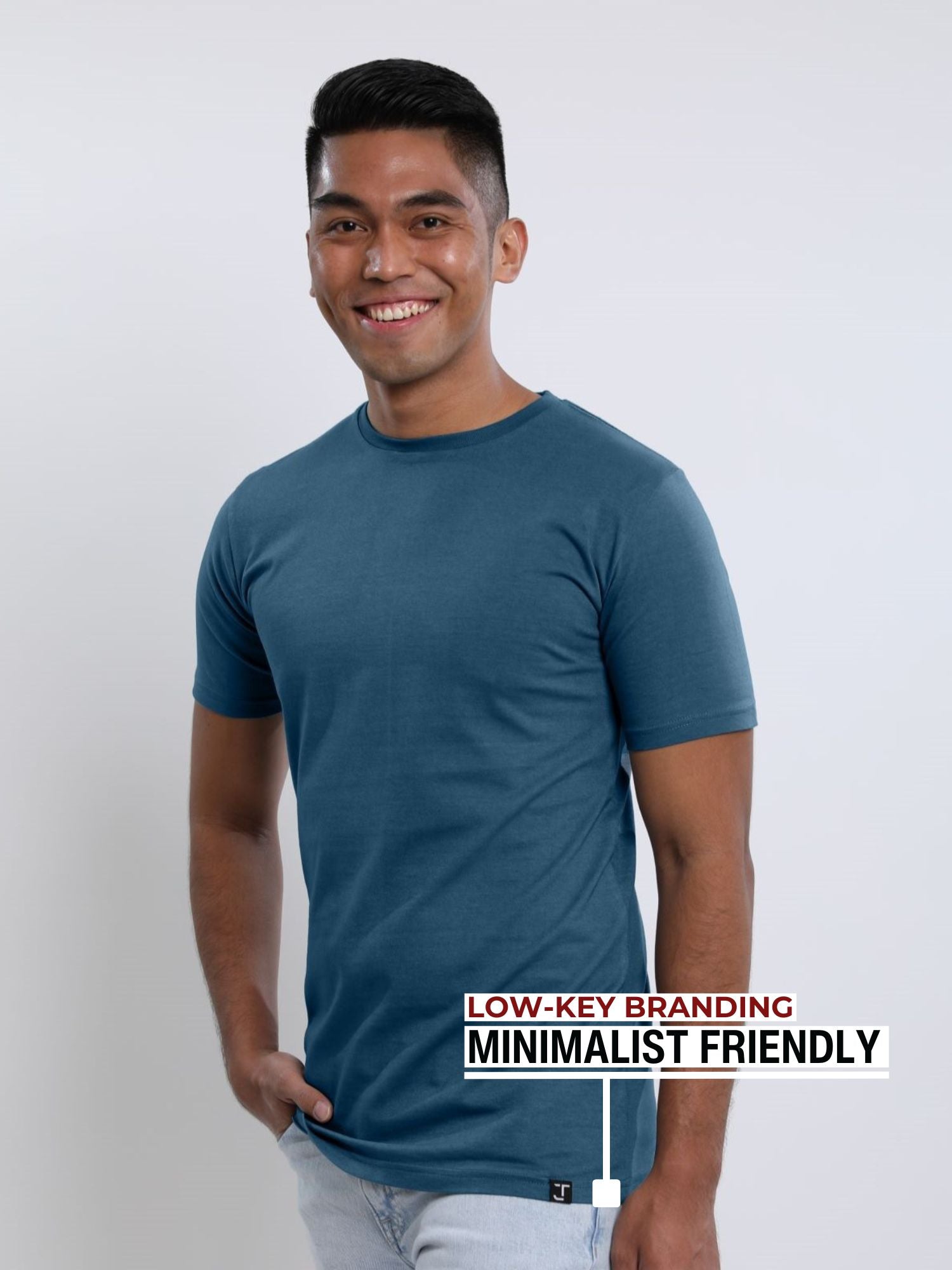 An upper body shot of a tall slim guy in a studio and wearing a petrol medium tall t-shirt, smiling and hand in pocket.