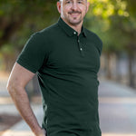 A tall and muscular man standing in a park, hands in back pockets and wearing a tall dark pine XL tall polo shirt.