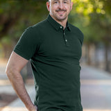 A tall and muscular man standing in a park, hands in back pockets and wearing a tall dark pine XL tall polo shirt.