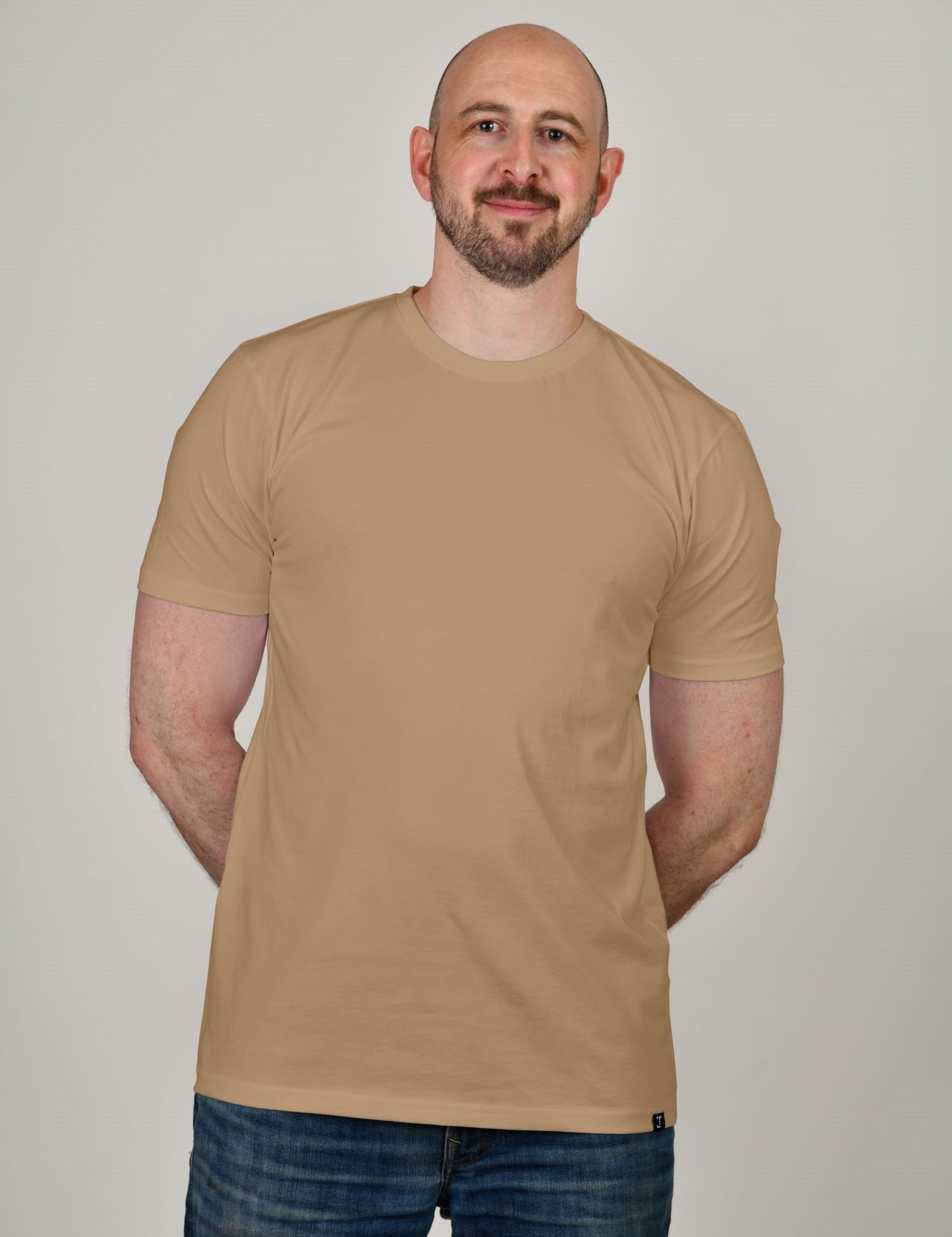 A tall and slim man in the studio standing in front of a light background. The smiling model is wearing an extra long slim tan t-shirt in a size XL. The tall tan t-shirt features a 3" longer body, 100% organic cotton, and is soft & preshrunk. The tan t-shirt is ideal for tall slim men 6'2"+.
