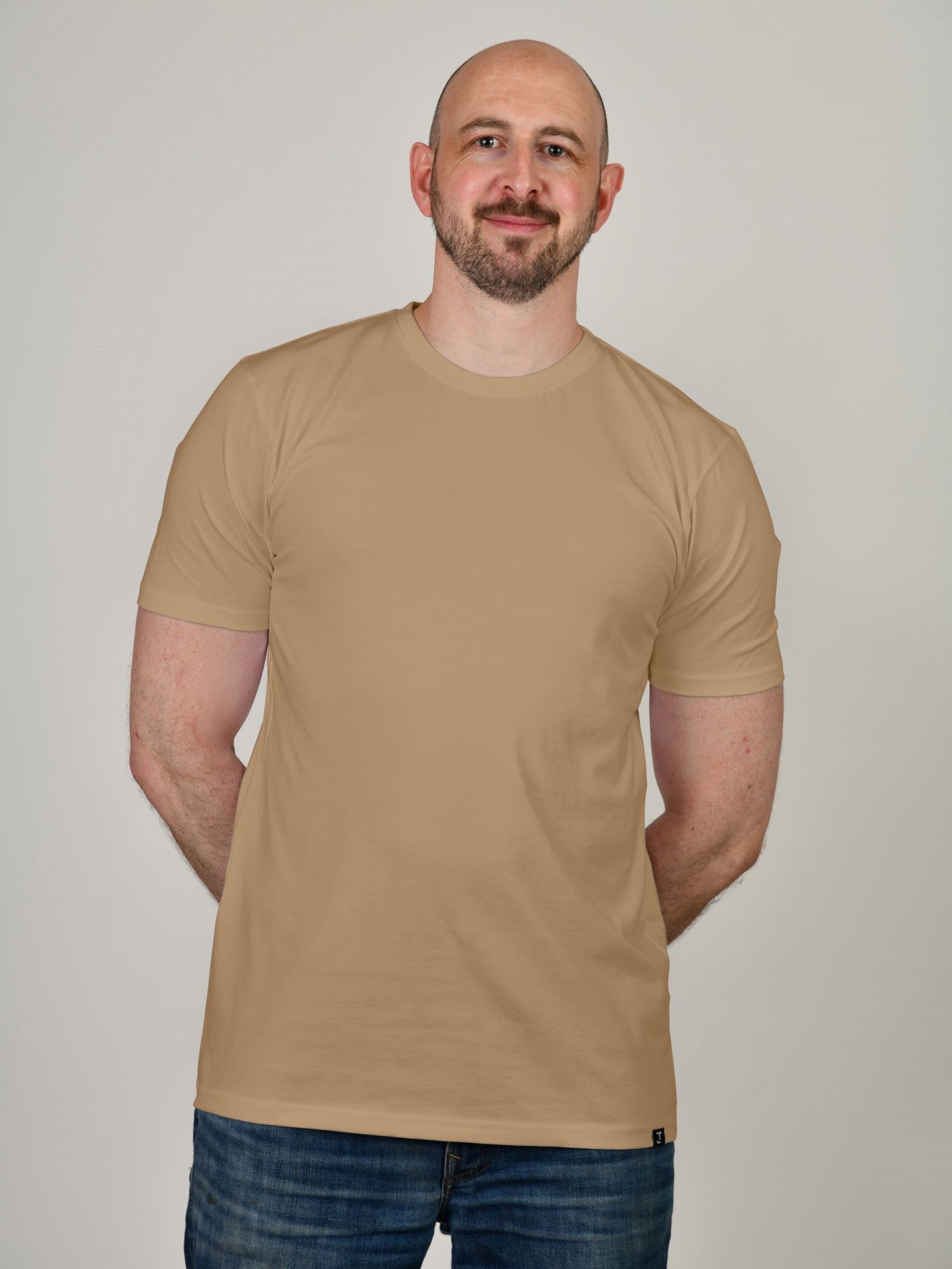 A tall and slim man in the studio standing in front of a light background. The smiling model is wearing an extra long slim tan t-shirt in a size XL. The tall tan t-shirt features a 3" longer body, 100% organic cotton, and is soft & preshrunk. The tan t-shirt is ideal for tall slim men 6'2"+.