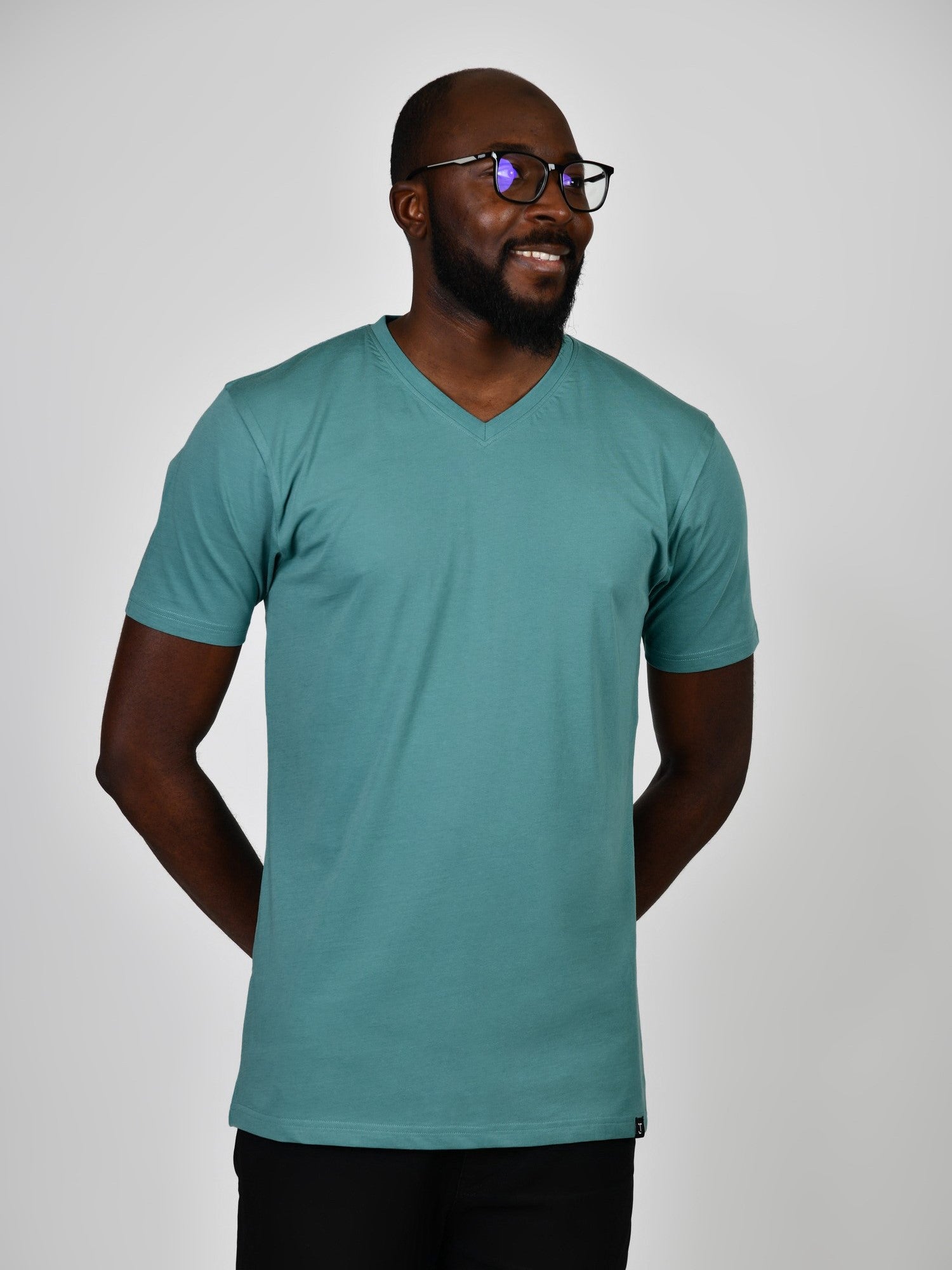 A tall and slim man in the studio standing in front of a light background with hands behind back. The smiling model is wearing an extra long slim teal v-neck t-shirt in a size large. The tall teal v-neck t-shirt features a 3" longer body, 100% organic cotton, and is soft & preshrunk. The teal v-neck t-shirt is ideal for tall slim men 6'2"+.