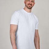 A tall and slim man in the studio standing in front of a light background. The smiling model is wearing an extra long slim white t-shirt in a size XL. The tall white t-shirt features a 3" longer body, 100% organic cotton, and is soft & preshrunk. The white t-shirt is ideal for tall slim men 6'2"+.