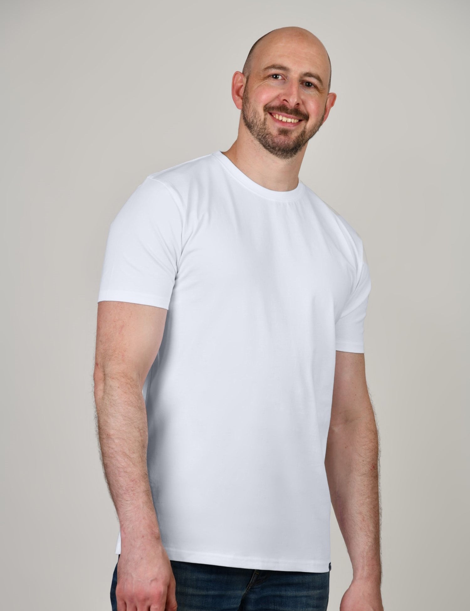 A tall and slim man in the studio standing in front of a light background. The smiling model is wearing an extra long slim white t-shirt in a size XL. The tall white t-shirt features a 3" longer body, 100% organic cotton, and is soft & preshrunk. The white t-shirt is ideal for tall slim men 6'2"+.