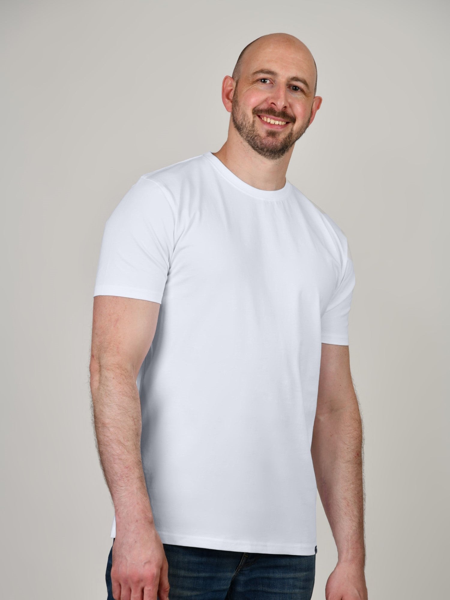 A tall and slim man in the studio standing in front of a light background. The smiling model is wearing an extra long slim white t-shirt in a size XL. The tall white t-shirt features a 3" longer body, 100% organic cotton, and is soft & preshrunk. The white t-shirt is ideal for tall slim men 6'2"+.