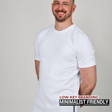 A tall athletic guy standing in a studio and wearing a white XL tall t-shirt, smiling and hands behind back.