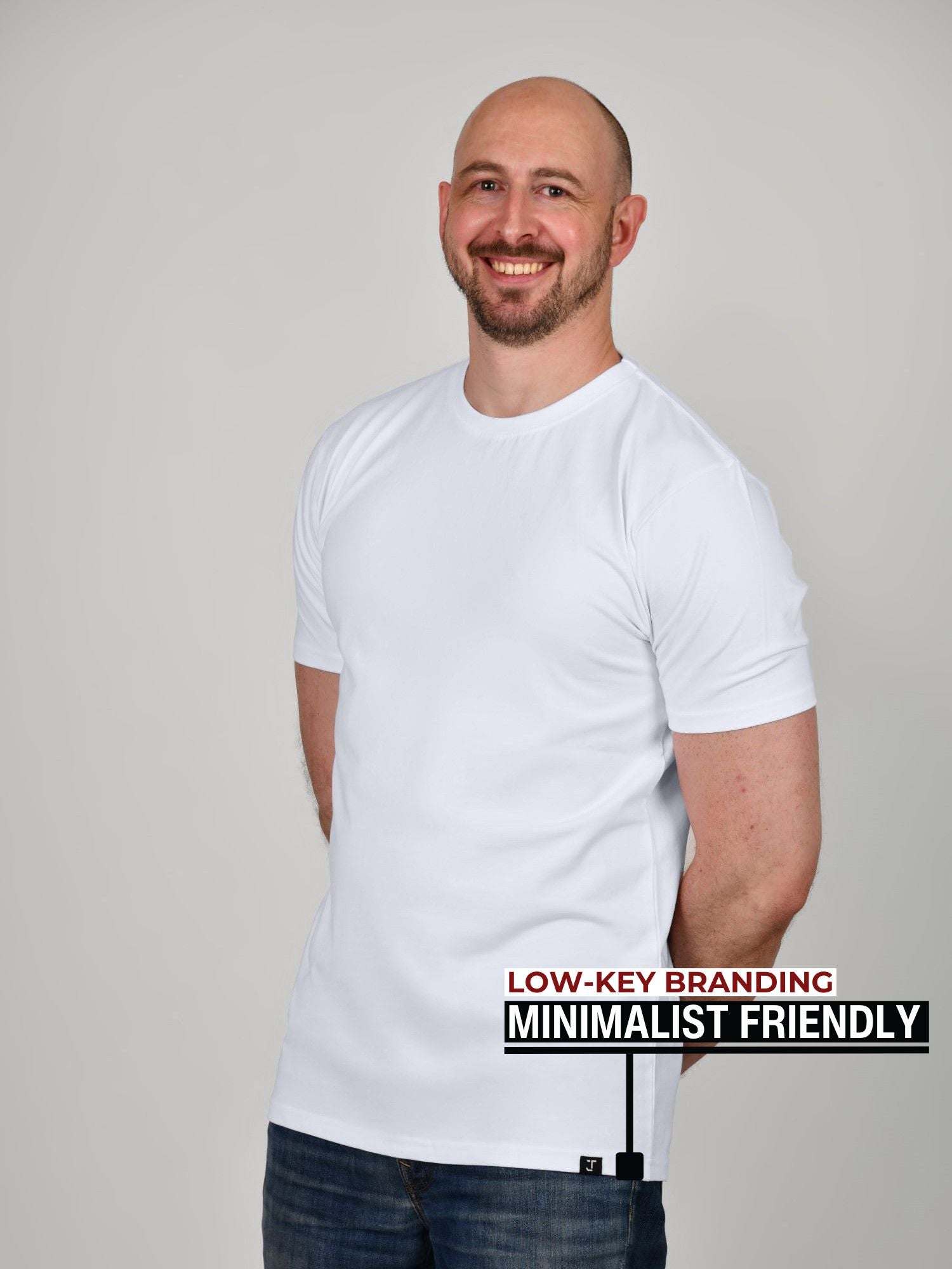 A tall athletic guy standing in a studio and wearing a white XL tall t-shirt, smiling and hands behind back.