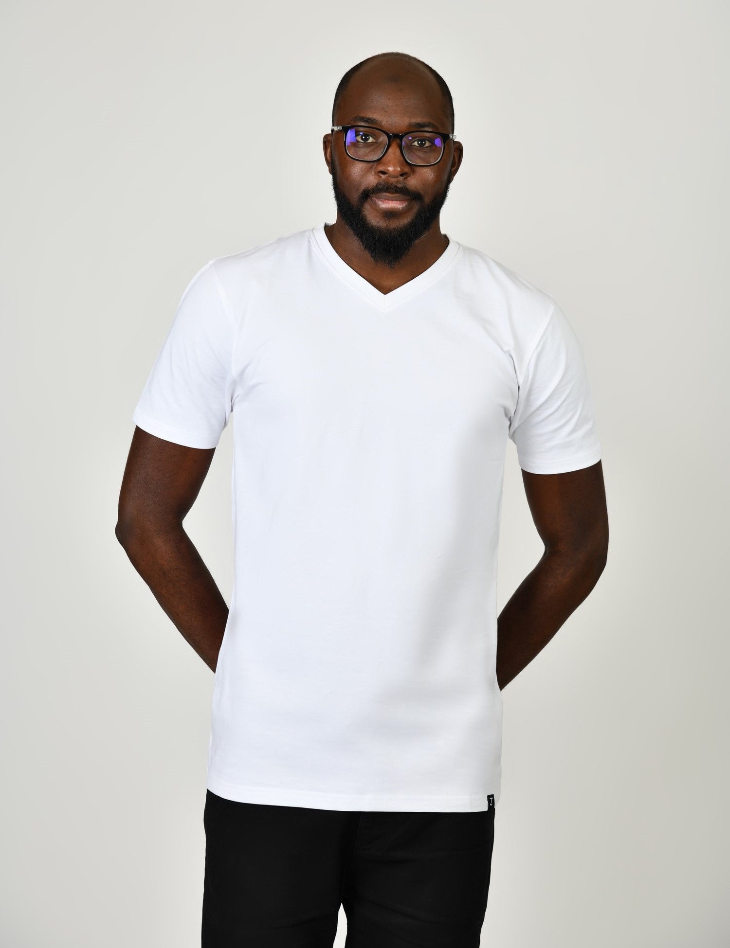 A tall and slim man in the studio standing in front of a light background with hands behind back. The model is wearing an extra long slim white v-neck t-shirt in a size large. The white v-neck t-shirt features a 3" longer body, 100% organic cotton, and is soft & preshrunk. The white v-neck t-shirt is ideal for tall slim men 6'2"+.