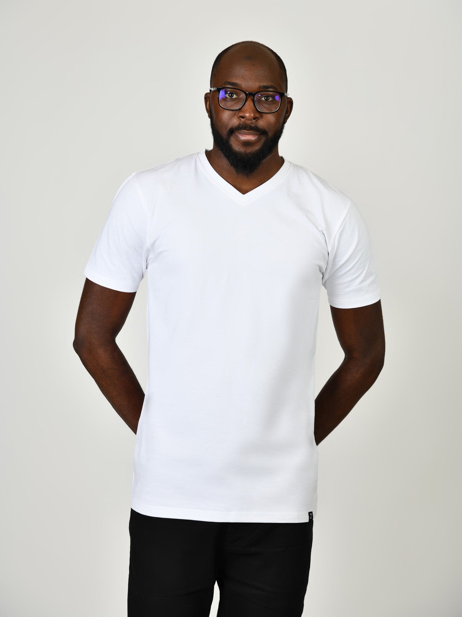 A tall and slim man in the studio standing in front of a light background with hands behind back. The model is wearing an extra long slim white v-neck t-shirt in a size large. The white v-neck t-shirt features a 3" longer body, 100% organic cotton, and is soft & preshrunk. The white v-neck t-shirt is ideal for tall slim men 6'2"+.