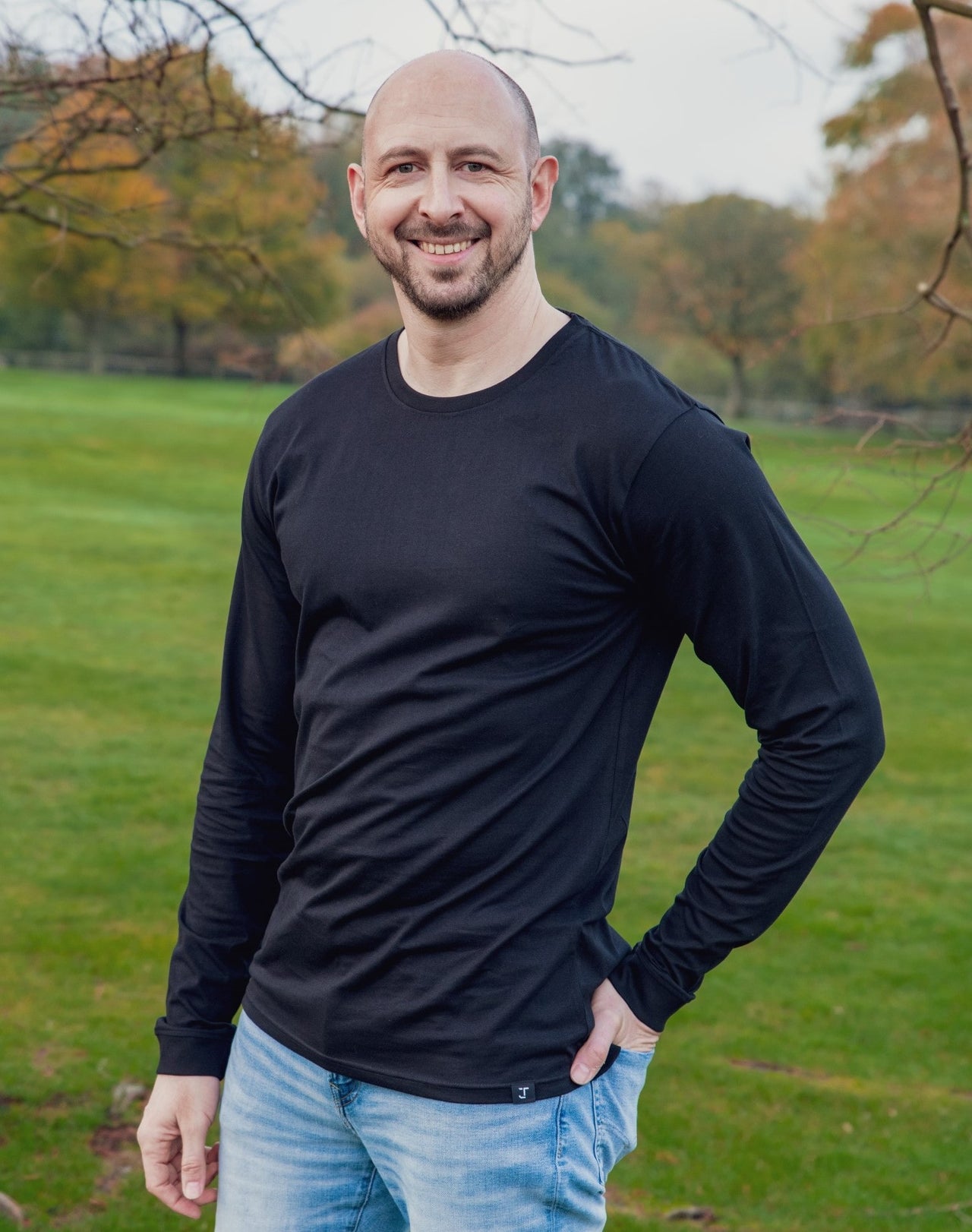 A tall athletic guy wearing a long sleeve black tall t-shirt and smiling in a park with hand in back pocket.
