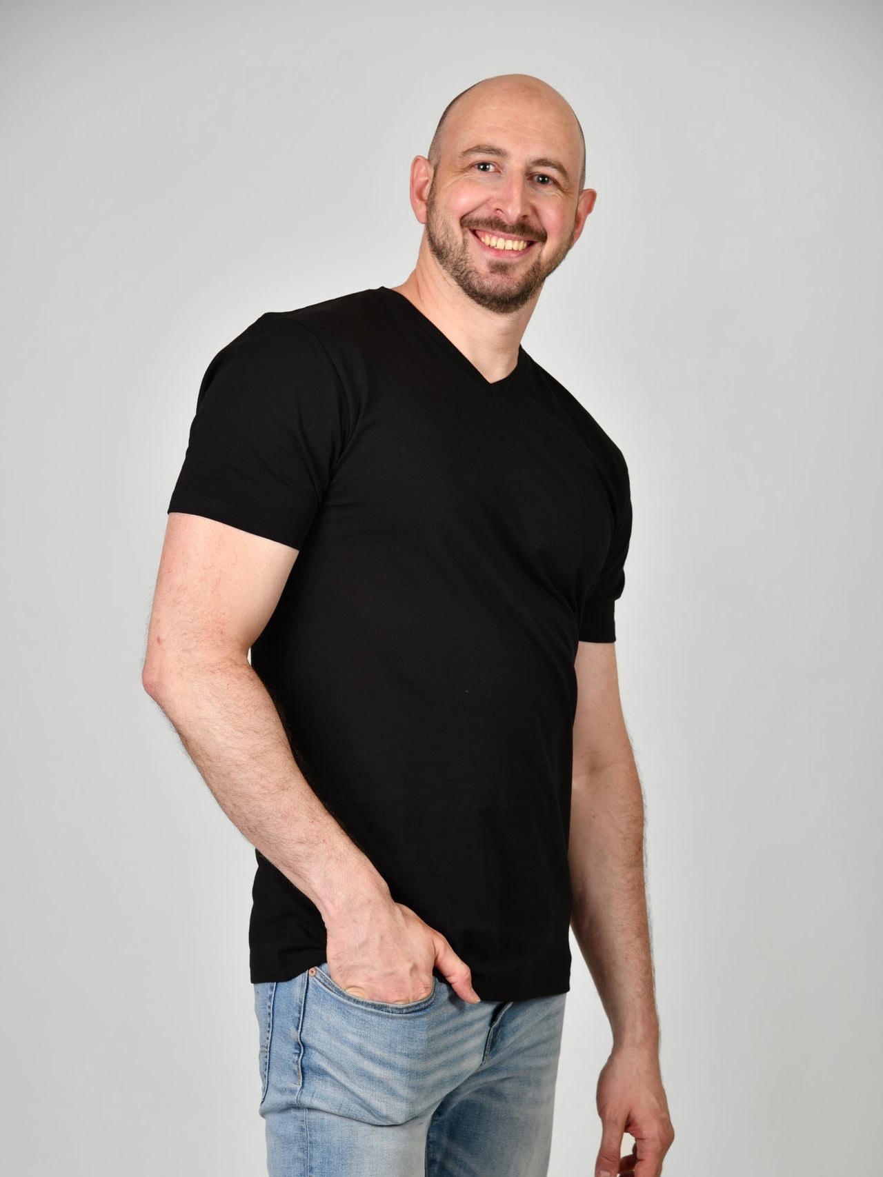 A tall and slim guy smiling in the studio, one hand in pocket and wearing a black XL tall slim v-neck t-shirt.
