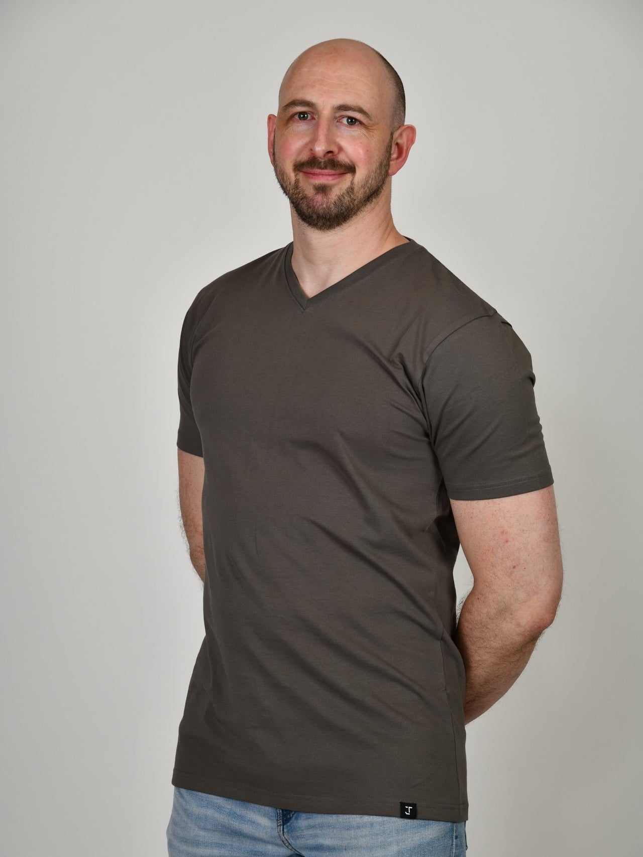 A tall and slim guy in the studio, hands behind back and wearing a dark grey XL tall slim v-neck t-shirt.