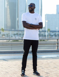Thumbnail for A head to toe shot of a tall and skinny guy in the street, arms folded, and wearing a white minimal graphic tall t-shirt.