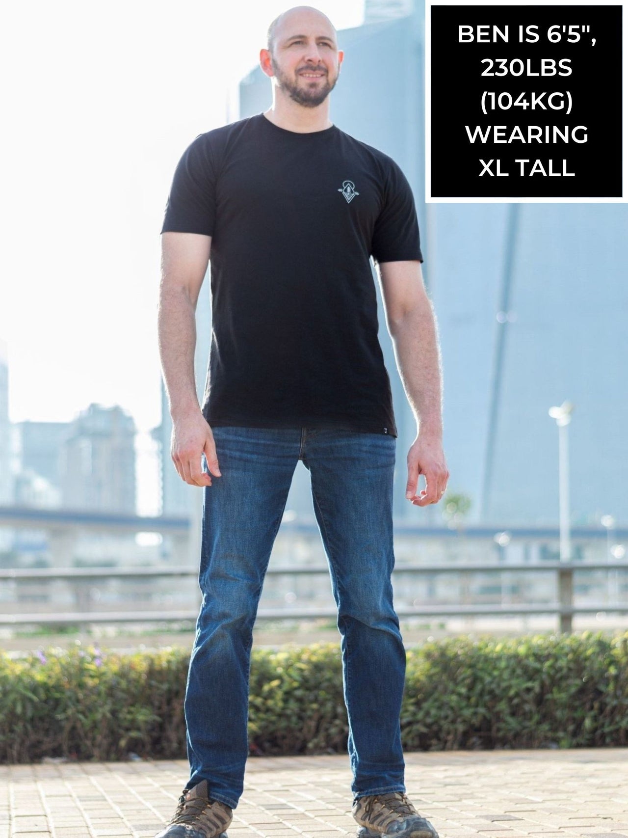 A head to toe shot of a tall and slim guy in the street and wearing a black minimal graphic tall t-shirt.