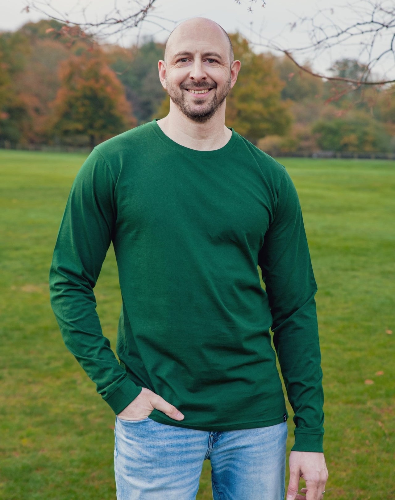 A tall athletic guy wearing a long sleeve dark green tall t-shirt and smiling in a park with one hand in pocket.