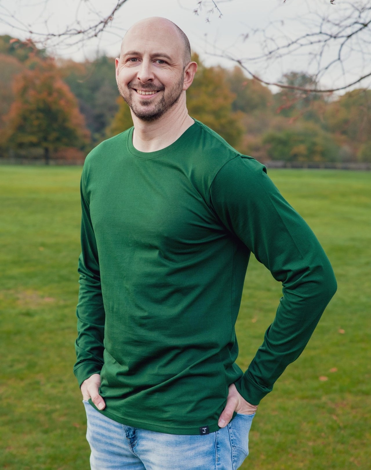 A tall athletic guy wearing a long sleeve dark green tall t-shirt and smiling in a park with hands in both pockets.