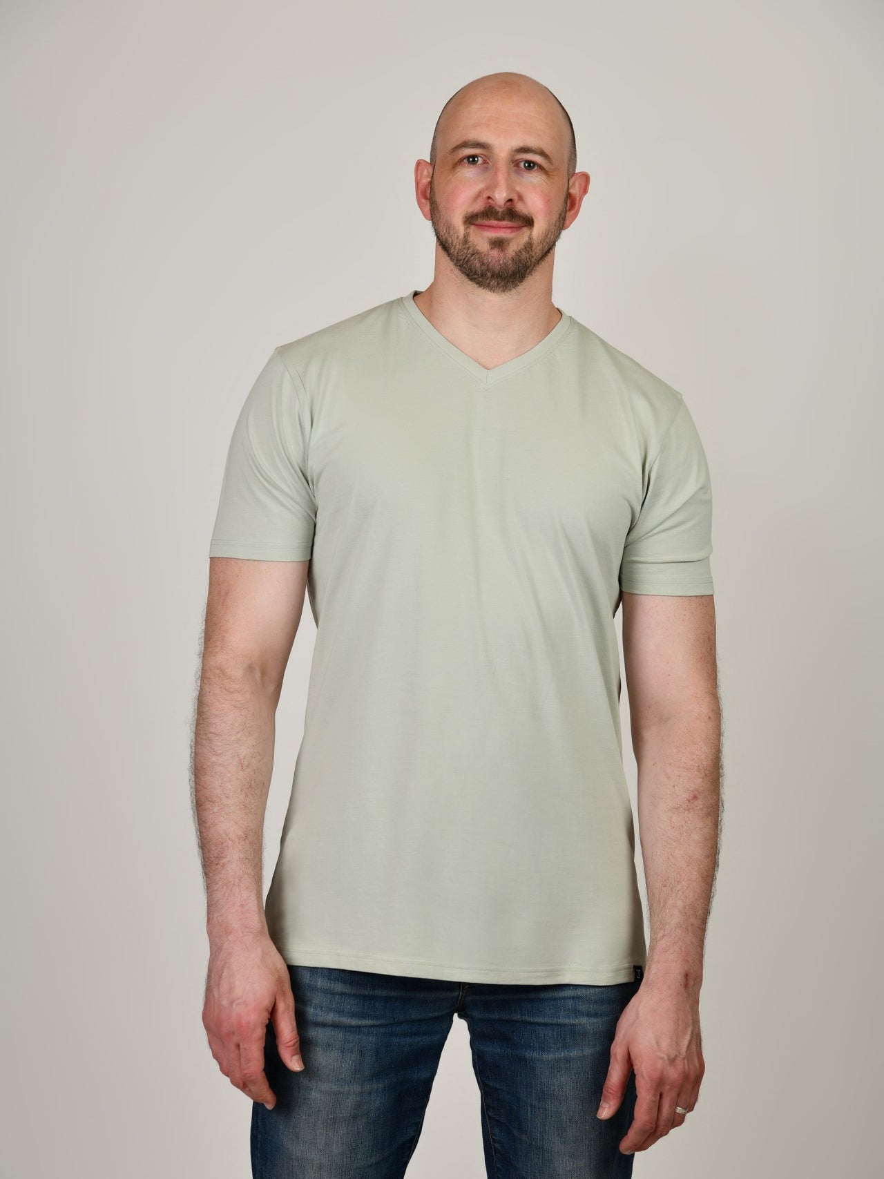 A tall and slim guy smiling in the studio and wearing a sage green XL tall slim v-neck t-shirt.