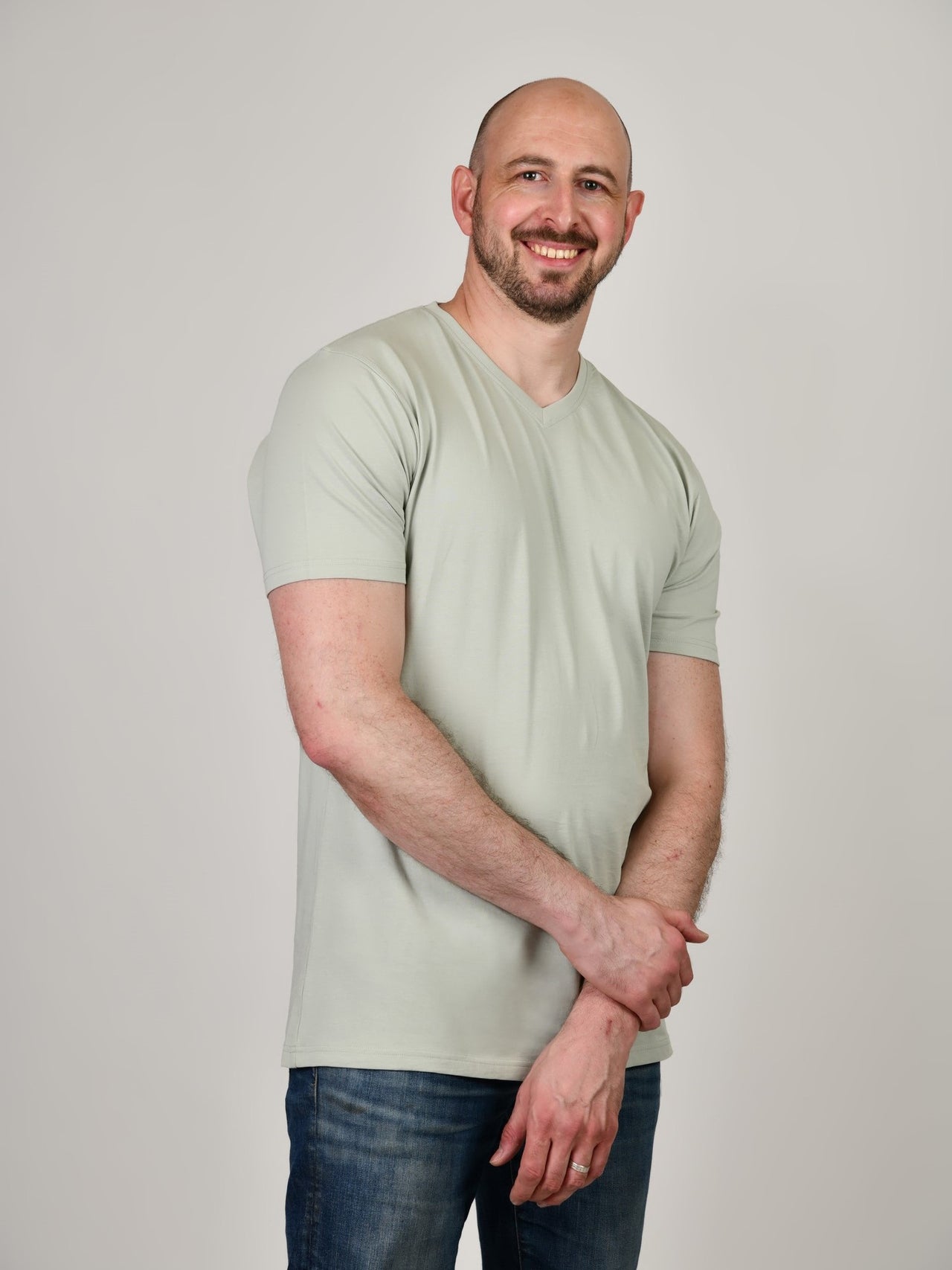 A tall and slim guy smiling in the studio, one hand grasping the other arm and wearing a sage green XL tall slim v-neck t-shirt.